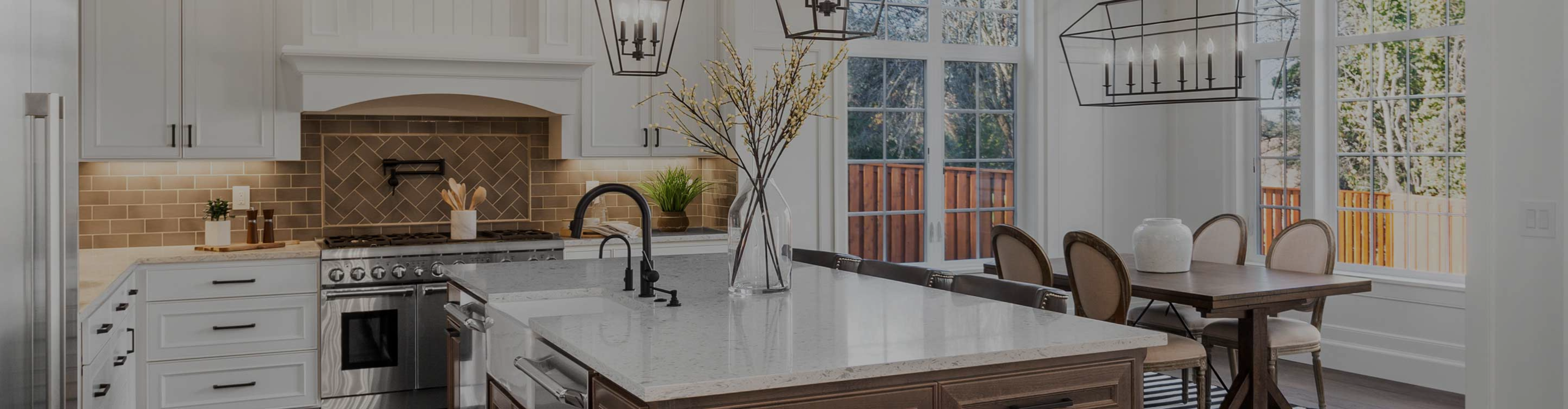 A modern kitchen with white cabinets with tile backsplash, a large island with a sink, and a dining area with a wooden table and chairs. Large windows overlook a backyard.